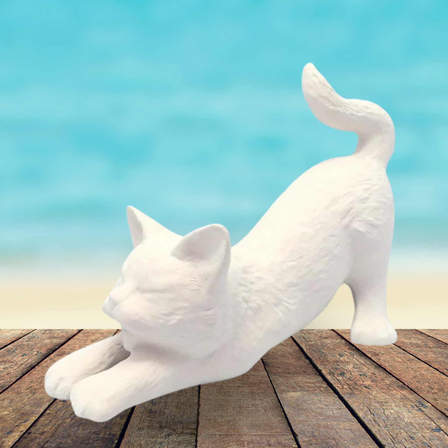 Handmade ready to paint ceramic cat figurine stretching in the downward dog position with its tail up. It is on a rustic wooden table by the ocean.