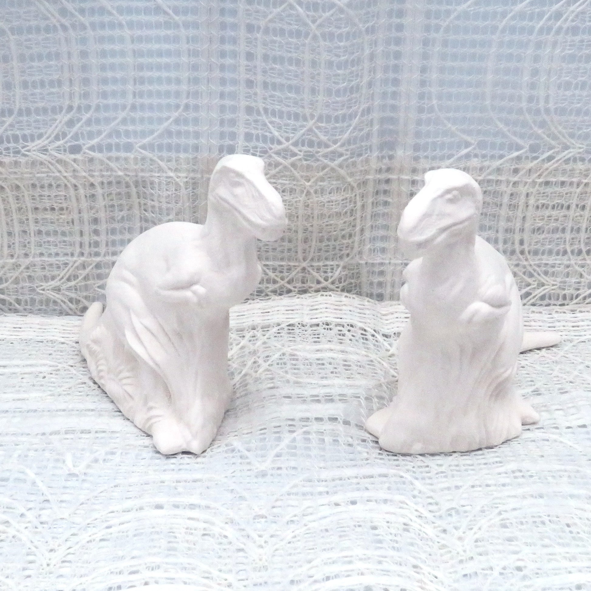 2 unpainted ceramic dino statues on a table with an ecru lacy  background