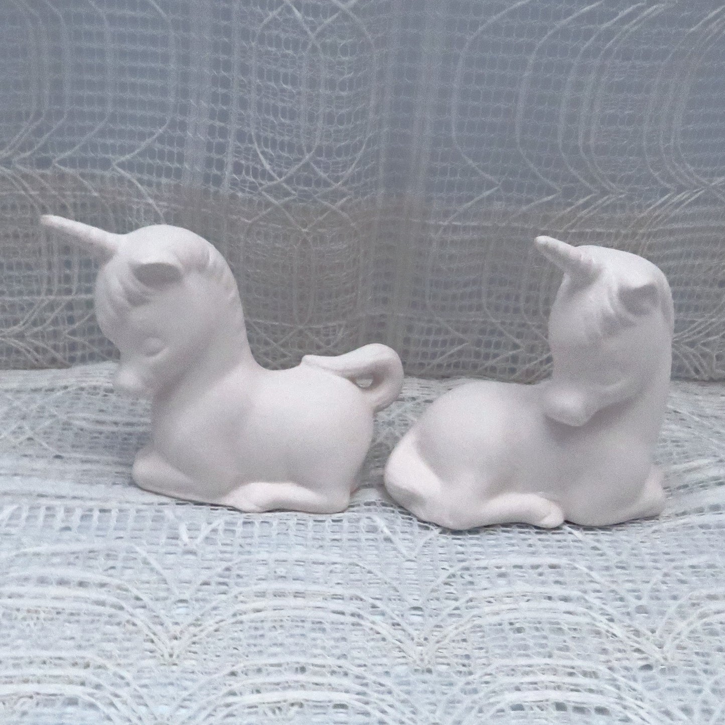 Handmade Ready to Paint Ceramic unicodrns lying tail to tail on a board with a lacy ecru curtain. One of the unicorns is looking backward at the first.