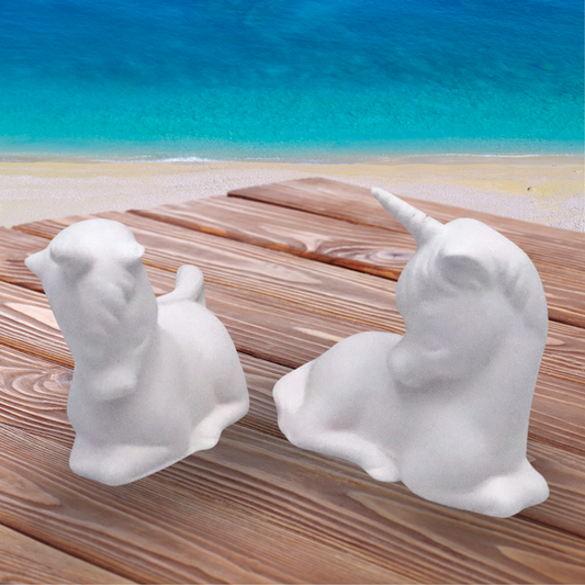set of 2 unpainted ceramic unicorn statues on a wood table by the beach.