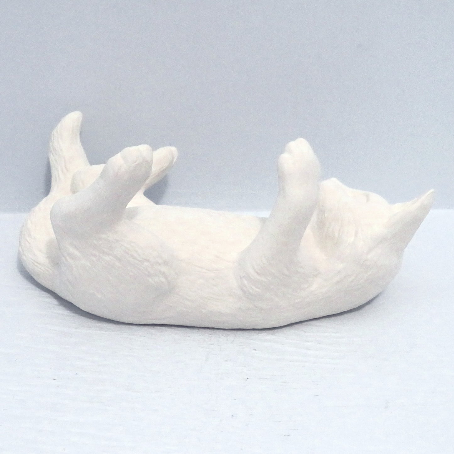Ready to Paint ceramic cat on back facing away from the camera. Its legs are in the air and it is on a pale blue surface.