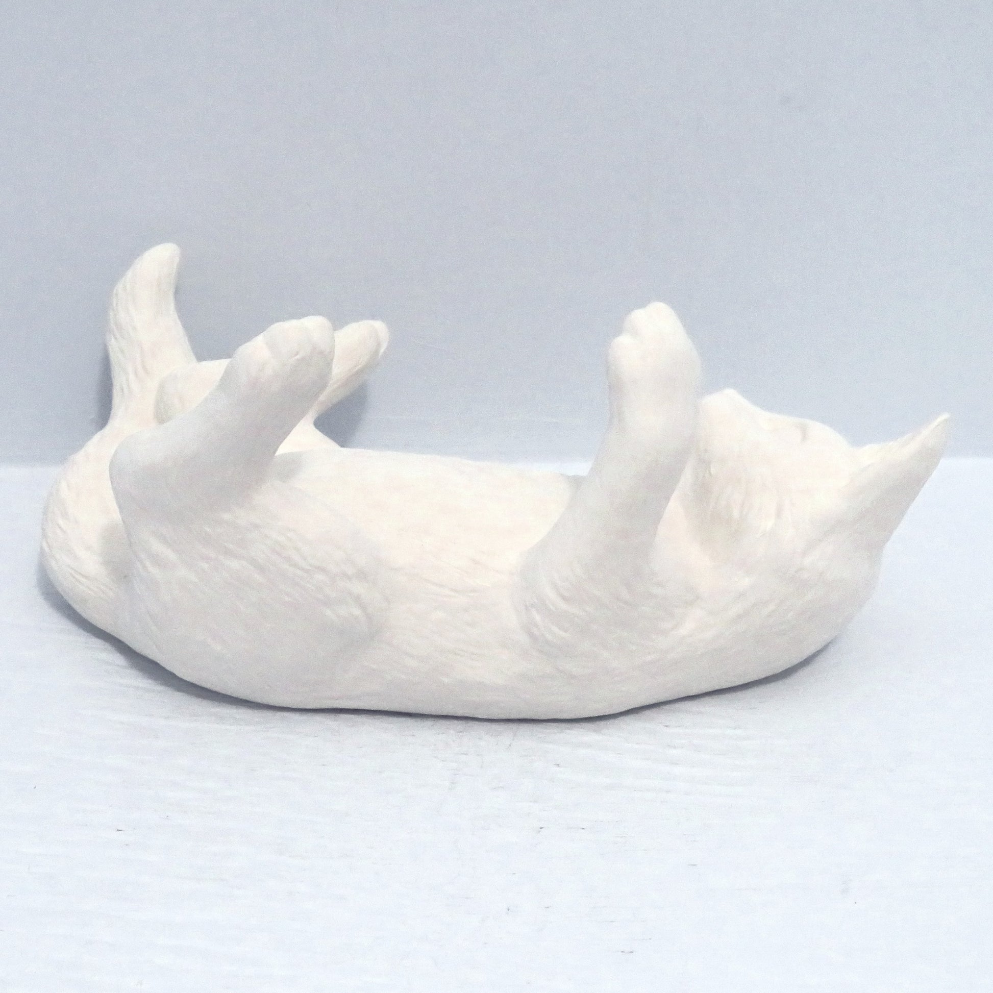 Ready to Paint ceramic cat on back facing away from the camera.  Its legs are in the air and it is on a pale blue surface.