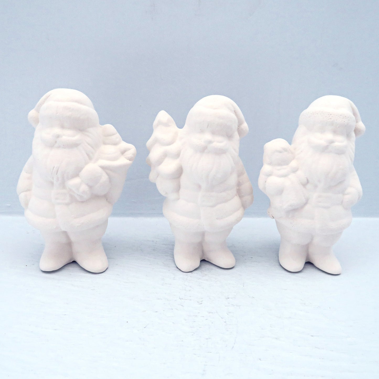 Set of 3 handmade ready to paint ceramic Santas. One is carrying a Christmas tree, one has a teddy bear, and one has other toys. They are facing forward on a pale blue surface.