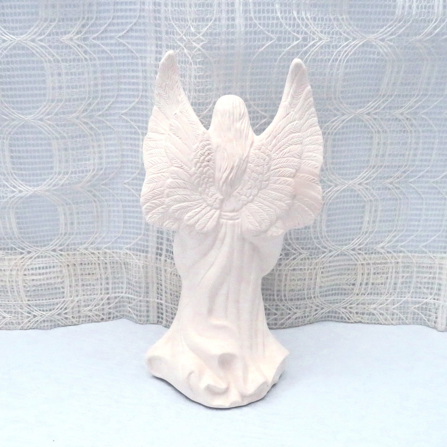 Rear view of paintable ceramic angel standing on a pale blue table with a lacy ecru curtain behind her.  You can see the detail in the wings and her hair.