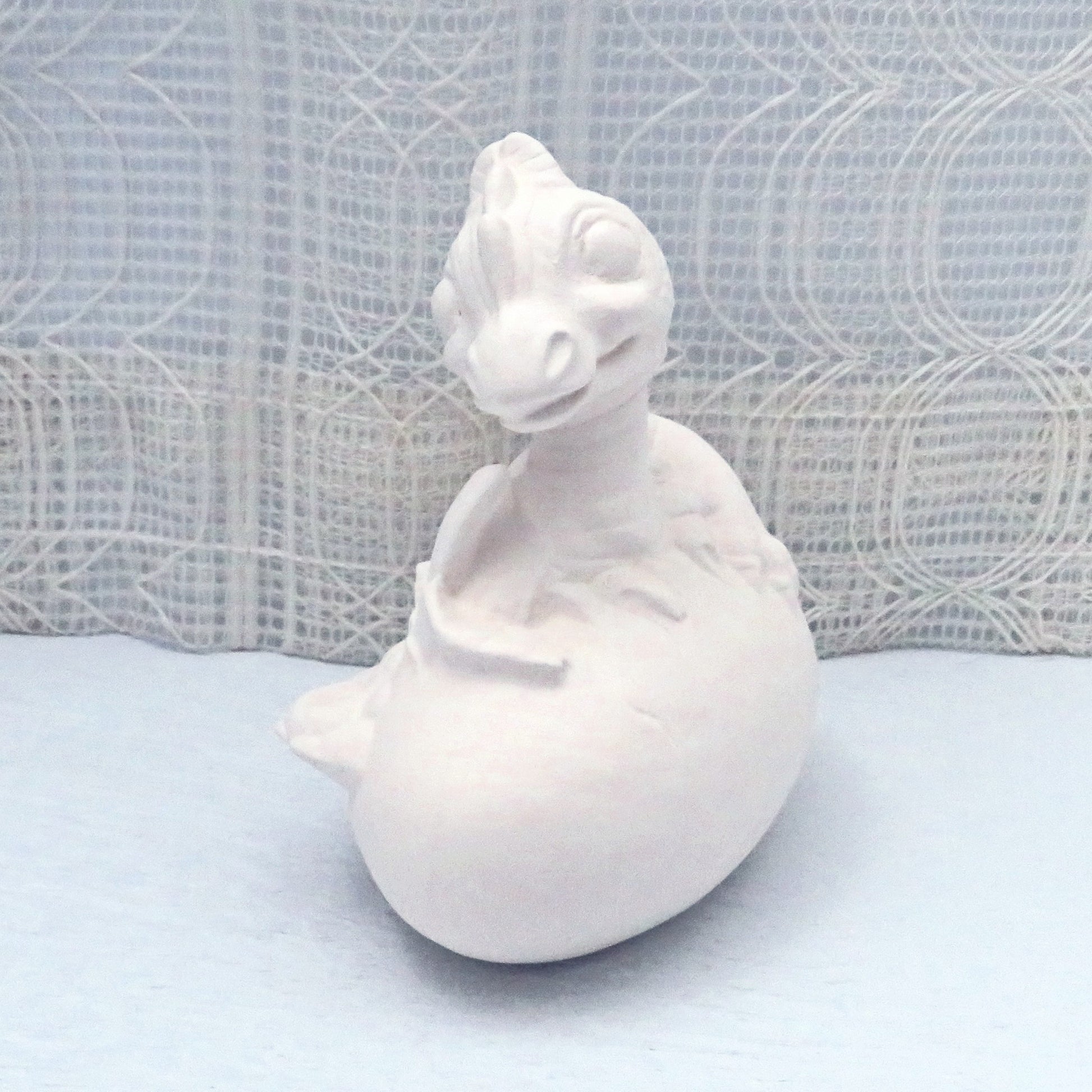 Side View of unpainted ceramic dragon coming out of her egg.  Her face is looking at the camera.