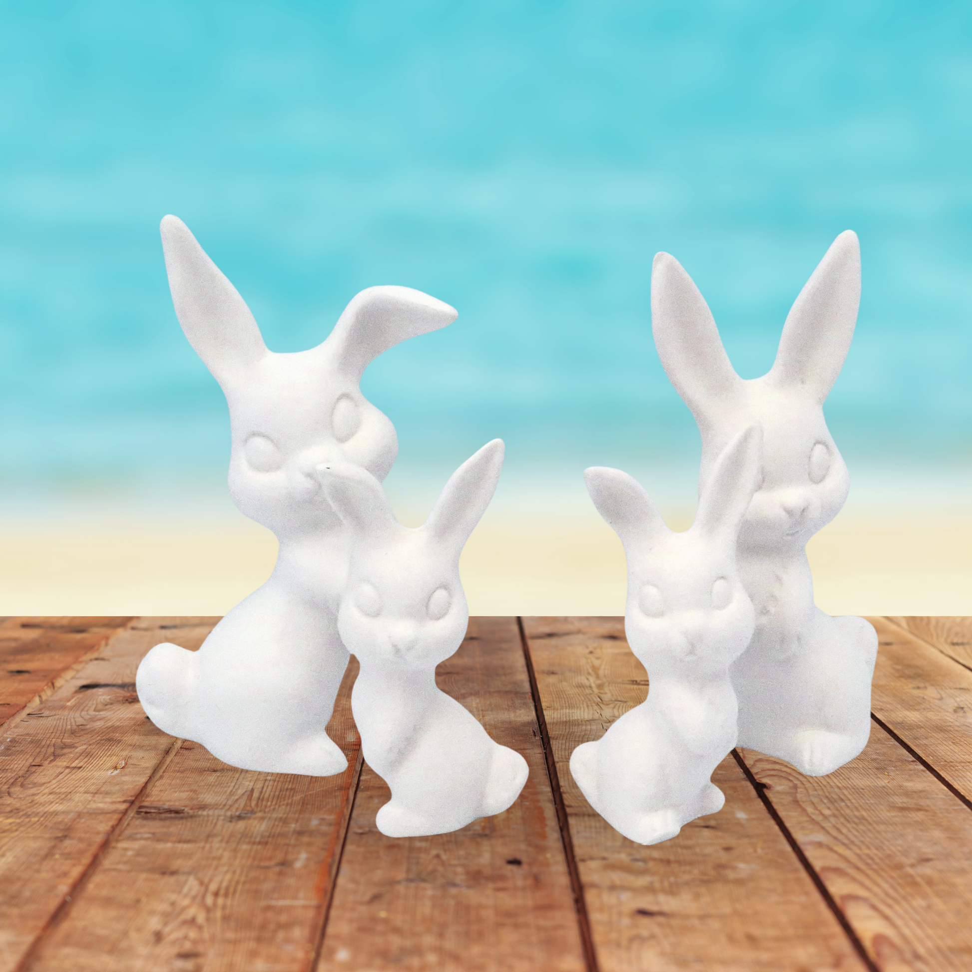 4 small ready to paint rabbit figurines standing on a rustic table with the ocean in the background.  There are 2 smaller ones, and 2 larger ones