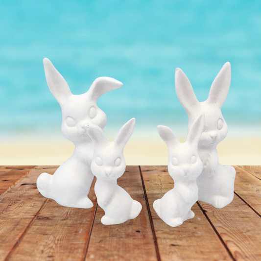 4 small ready to paint rabbit figurines standing on a rustic table with the ocean in the background.  There are 2 smaller ones, and 2 larger ones