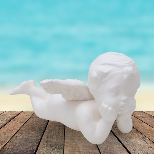 Unpainted cherub lying on his stomach with his head in his hands.  He is on a rustic wood table by the ocean.