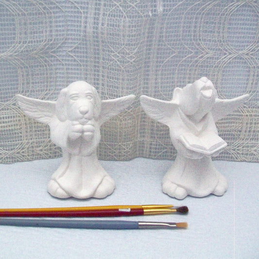 2 ready to paint dog angel figurines on a pale blue table with an ecru curtain behind them.  One is holding a book and howling, one appears to be praying.  There are 3 paint brushes in the foreground.