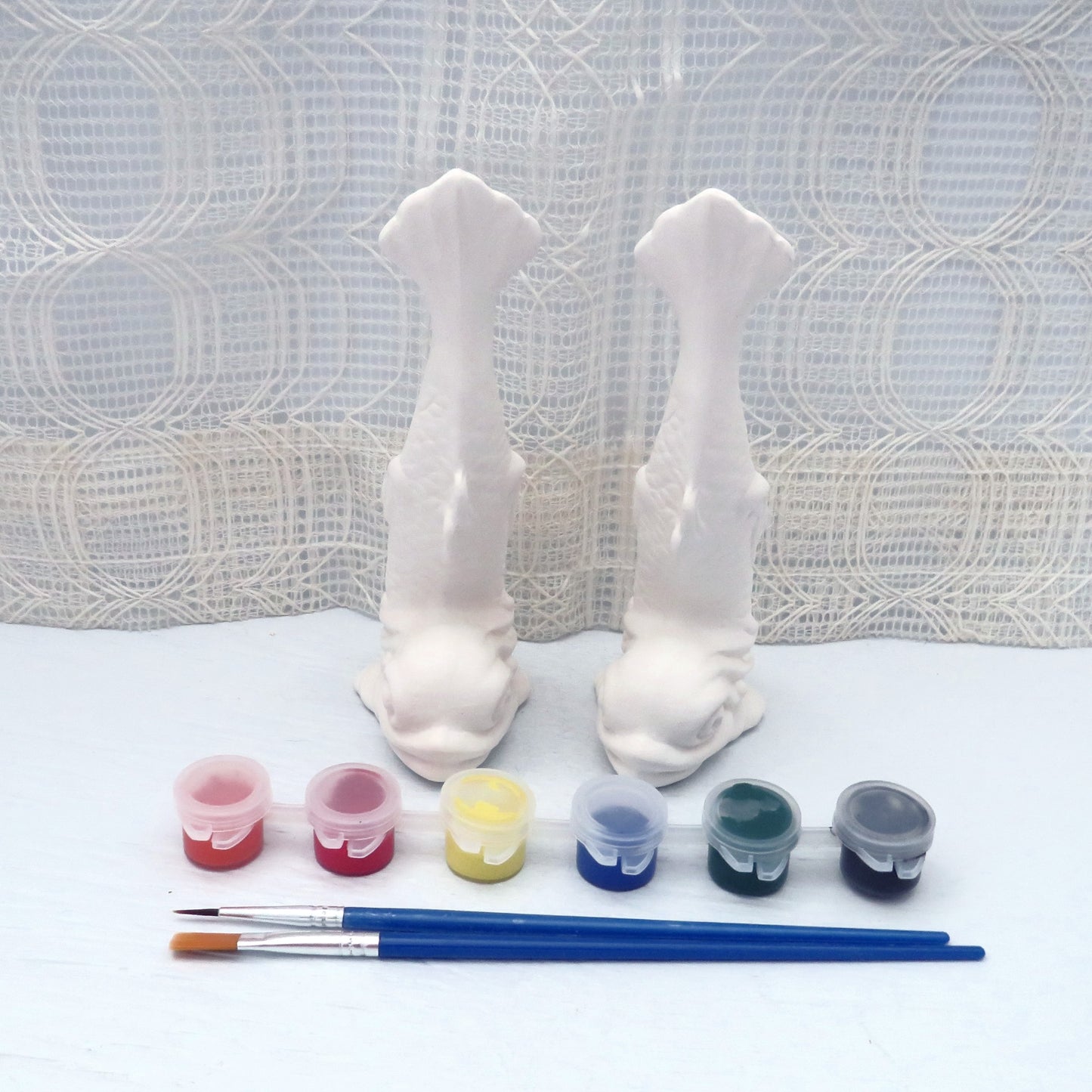 set of 2 ready to paint ceramic fish figurines with their tails up sitting on a pale blue table with an ecru lacy curtain behind them.  There is a set of 6 acrylic paints and 2 paint brushes in front of them.