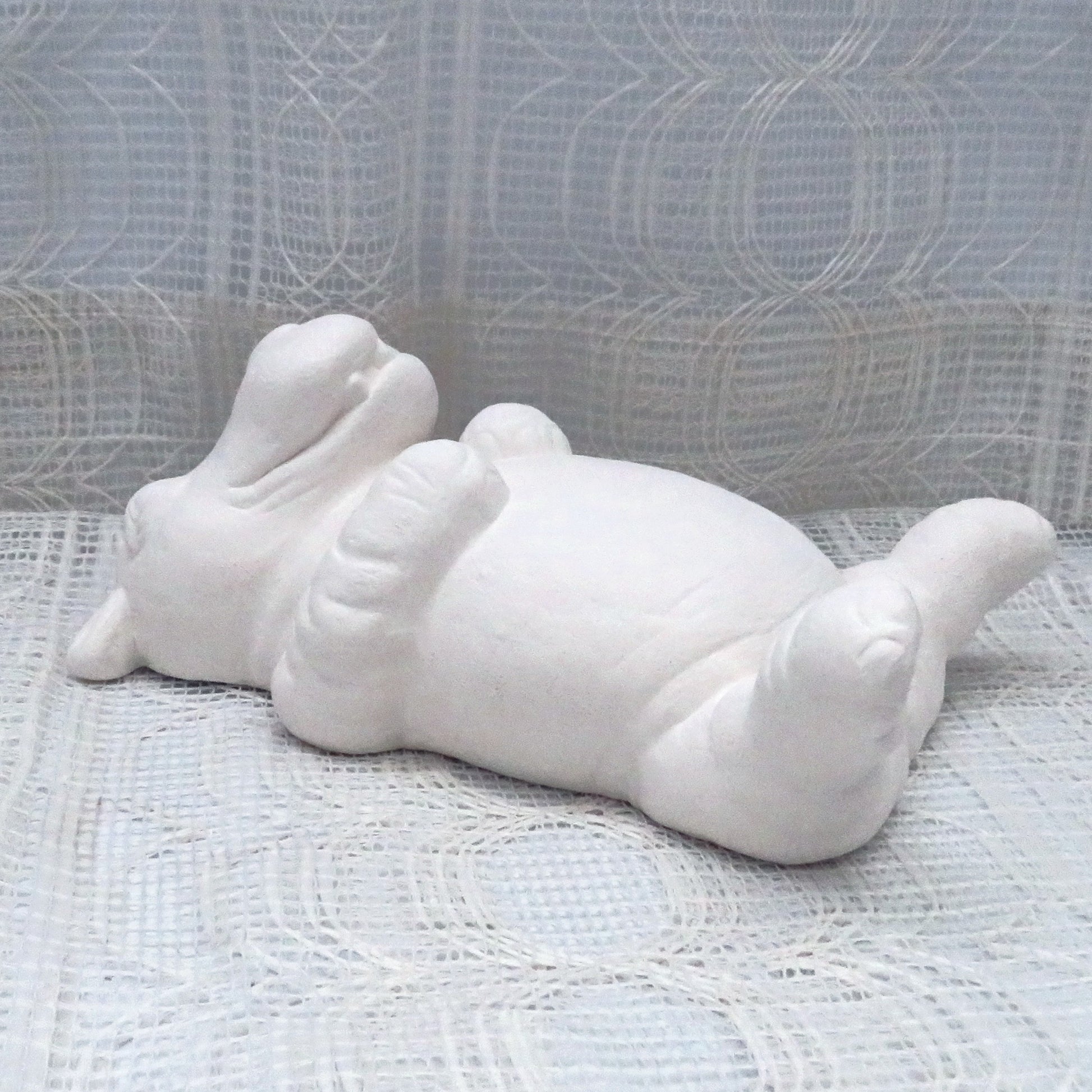 Ready to Paint ceramic hippo lying on back with arms across his chest and rear legs in hair.  It has a silly grin on his face and is lying on a table with an ecru lacy table cloth.l