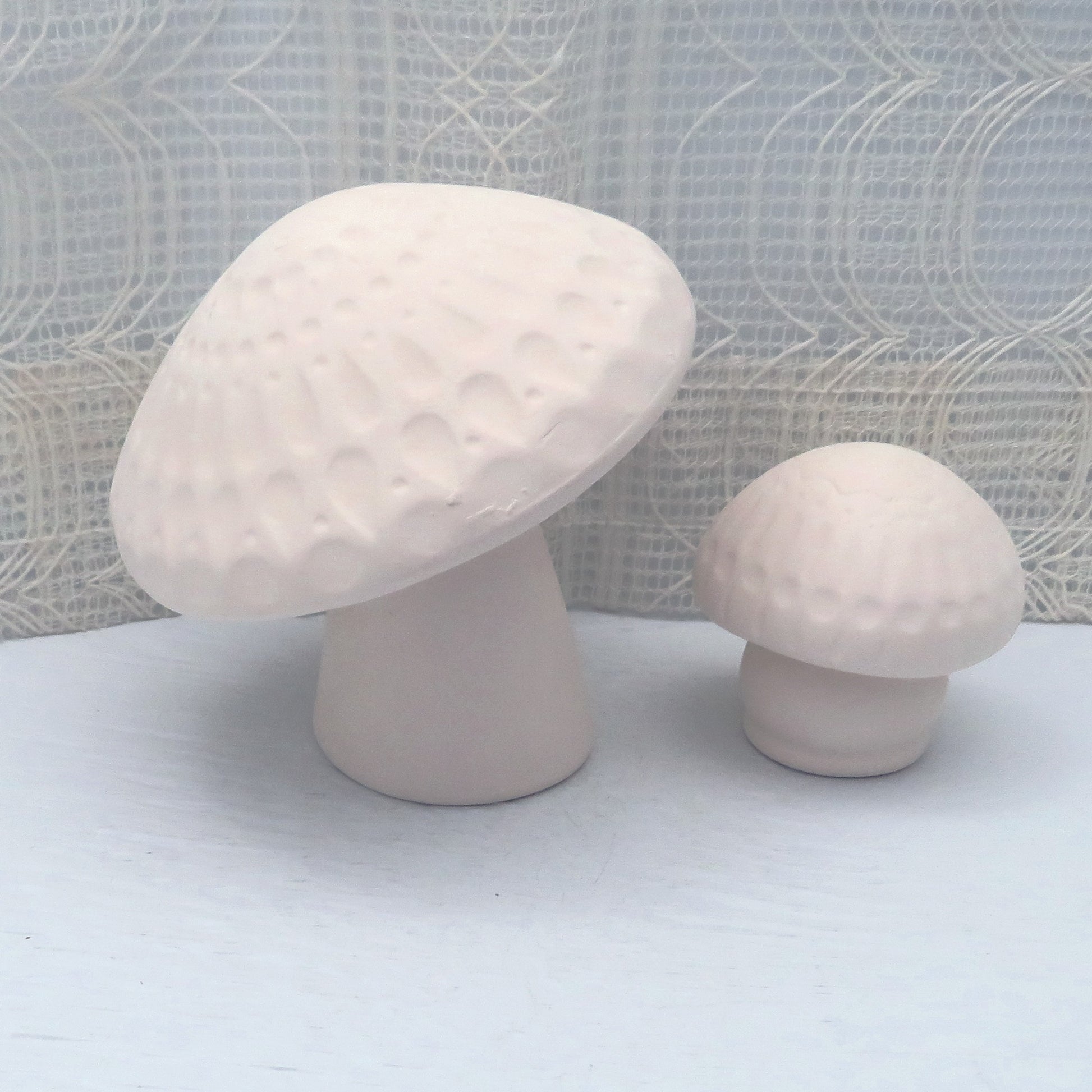 Paintable ceramic mushroom statues on a blue table with an ecru lacy curtain behind it.  You can see and feel the texture in the tops of the mushrooms.  One mushroom is larger, one smaller.
