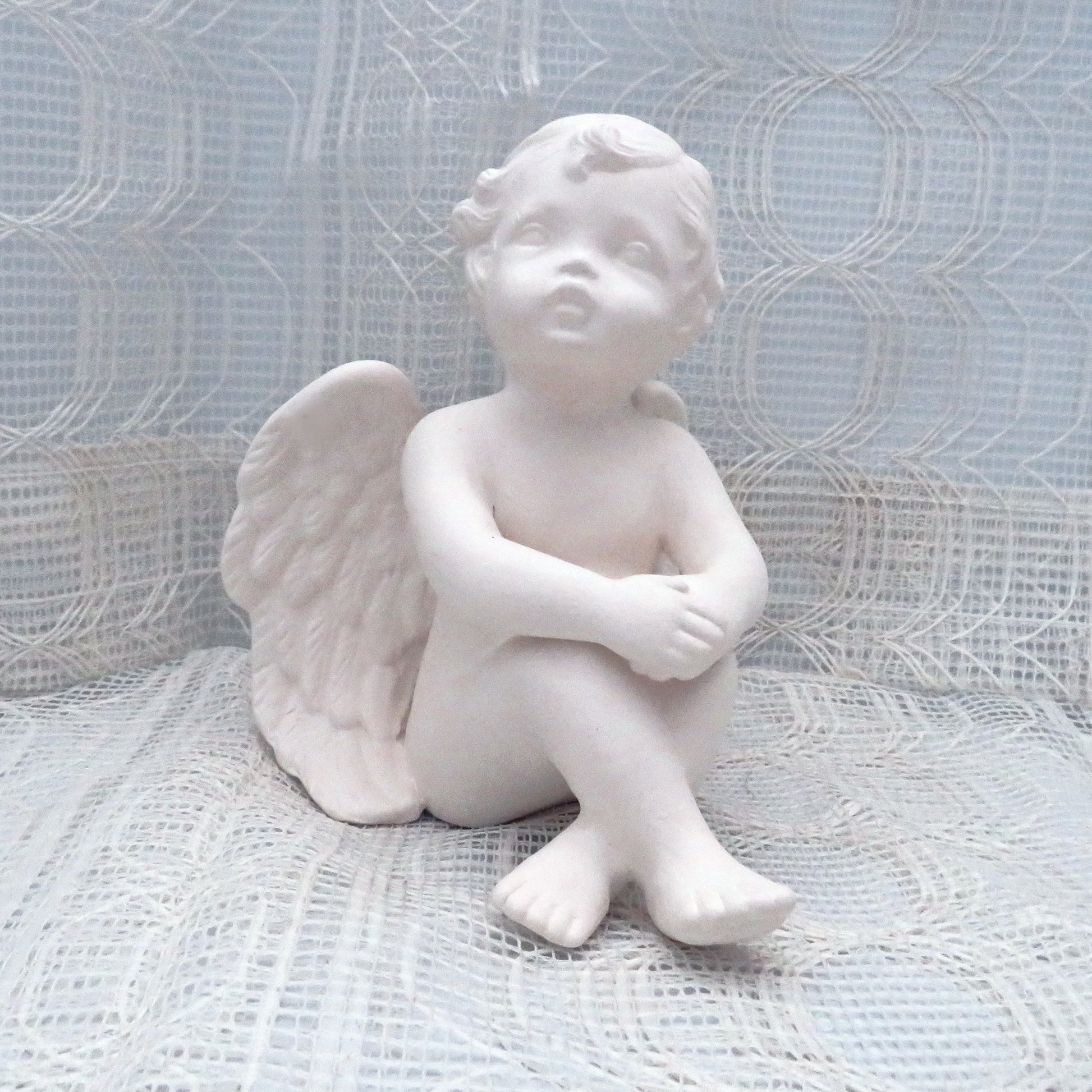 6 inch paintable ceramic cherub statue sitting on a lacy ecru table cloth with a curtain behind it.  His knees are up and his hands are resting on his knees.