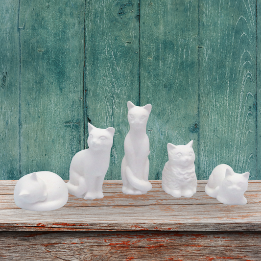 Five white unpainted ceramic cat figurines in various poses, including sleeping, sitting, and standing, arranged on a rustic wooden surface against a distressed teal-green wooden plank background.