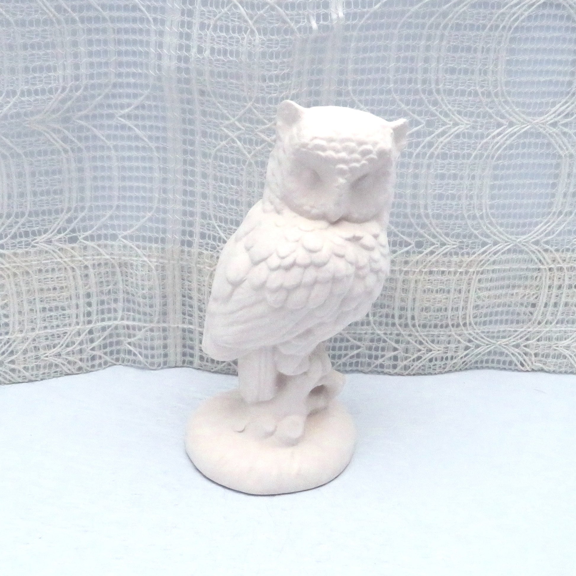 Ready to paint ceramic owl figurine sitting on a pale blue table with an ecru lacy curtain behind.  The owl is and looks like it si standing on a tree stump.