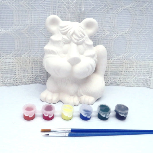 Ready to paint ceramic tiger figurine on a pale blue table with an ecru curtain beind it.  There is aset of 6 actylic paint pots and 2 paint brushes in front of it.