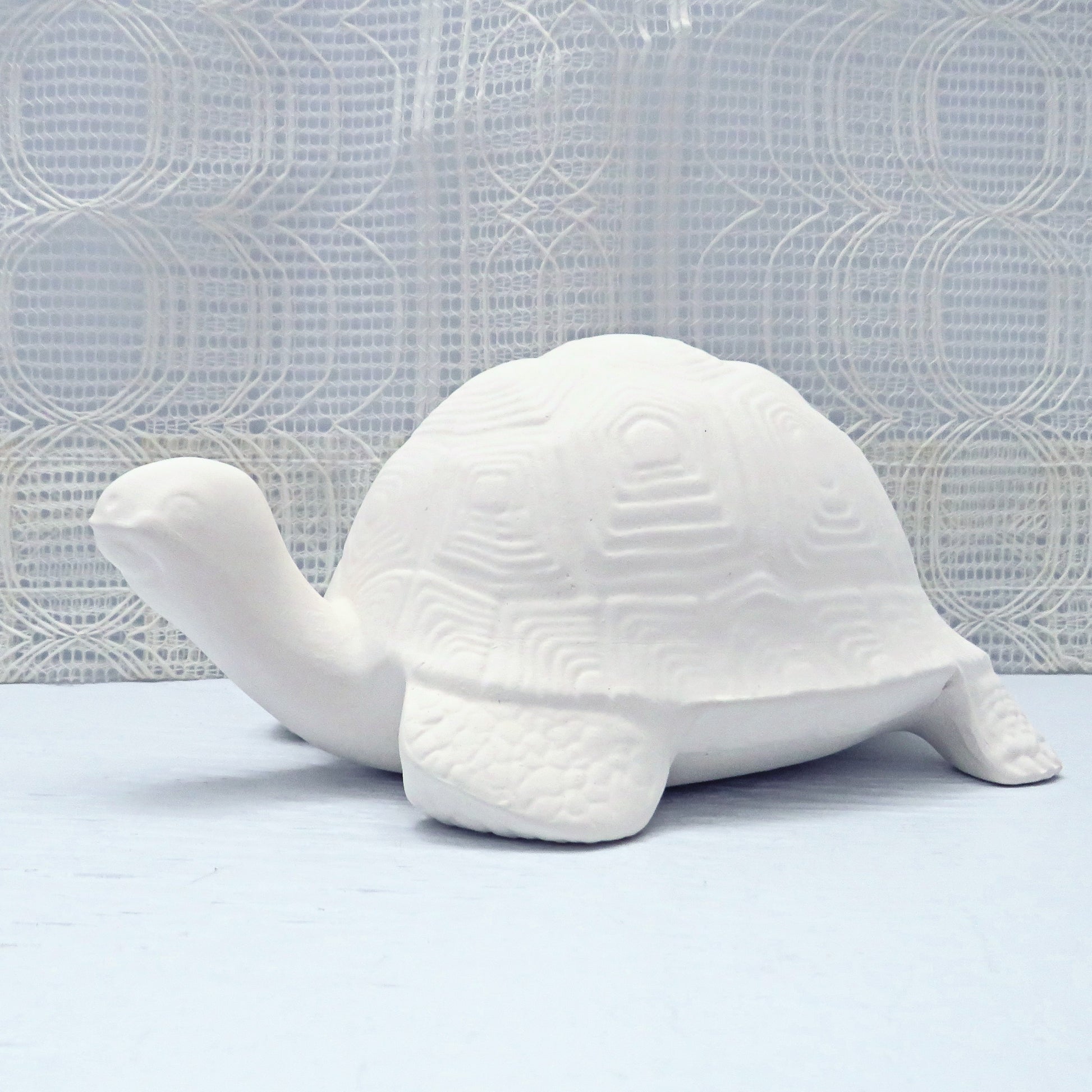 Unpainted ceramic turtle on a blue table with a lacy curtain.  the turtle is facing to the viewer's left