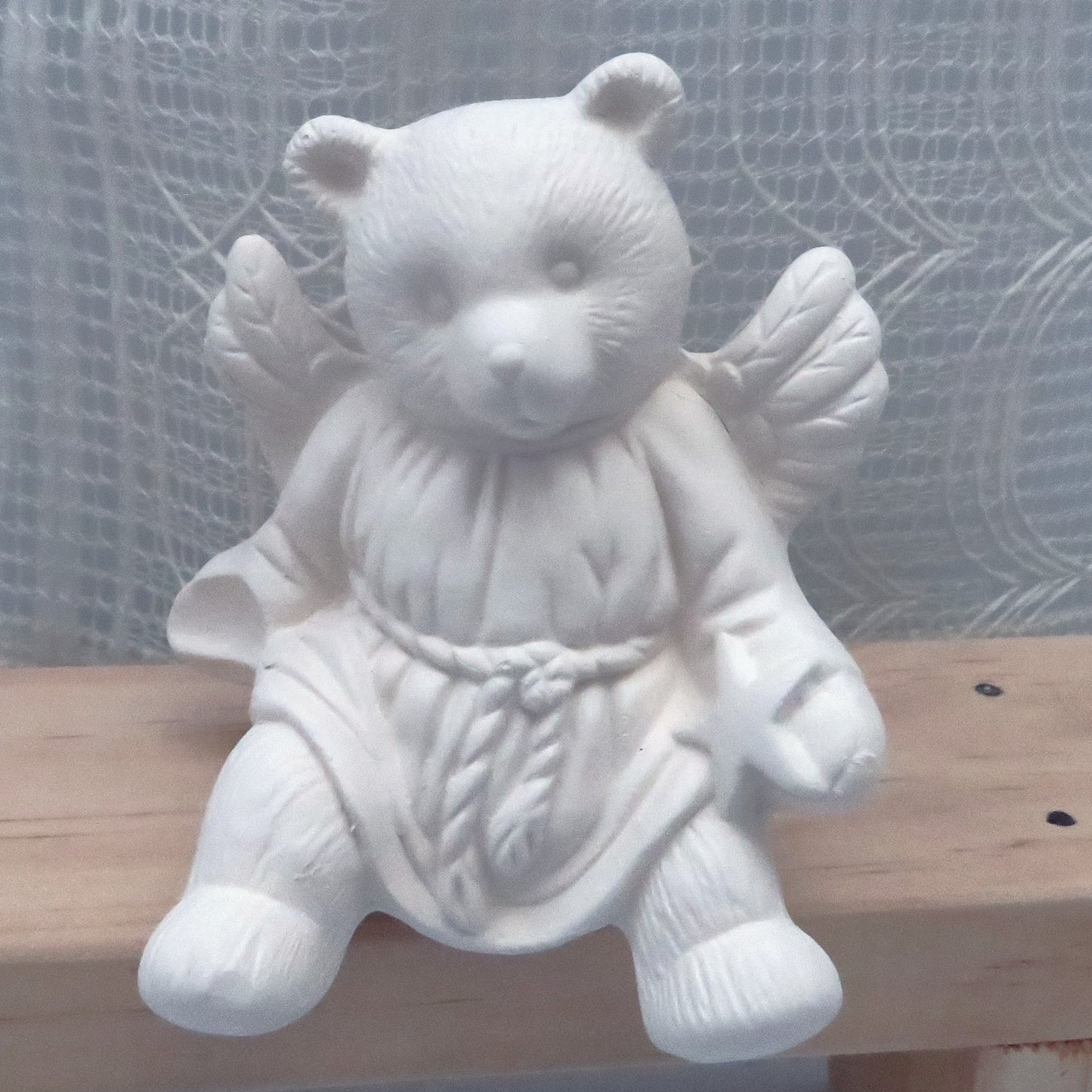 Close up view of unpainted ceramic bear angel sitting on a wooden bench.