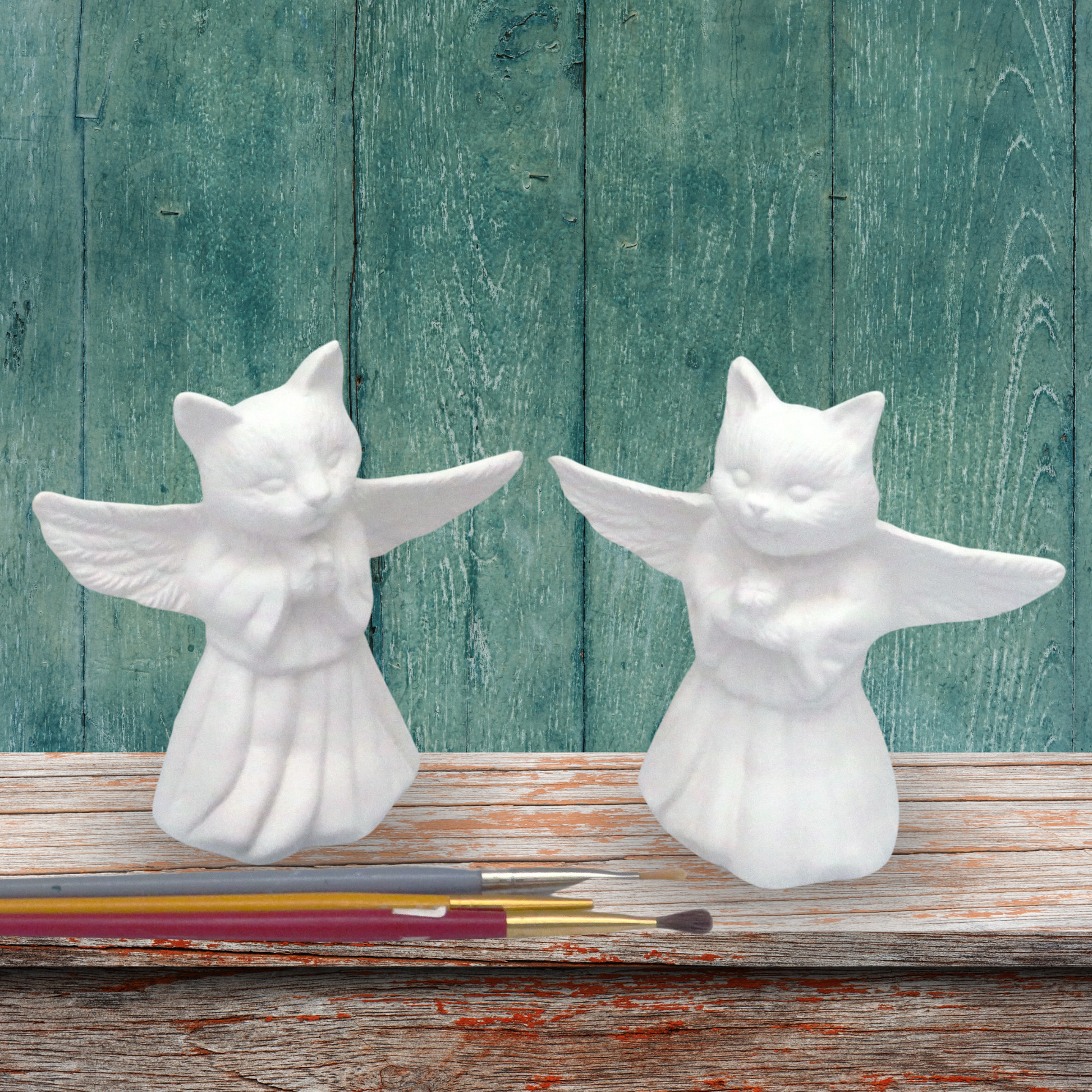 set of 2 ready to paint ceramic cat angels on a rustic wooden shelf with a green backgraound and 2 paint brushes