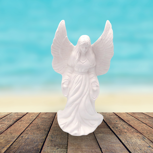 Handmade Ready to Paint ceramic angel statue standing on rustic brown wooden table by a smooth ocean background.  Her wings are outstretched, her head is slightly bowed.  Her arms are by her side and hands facing forward.