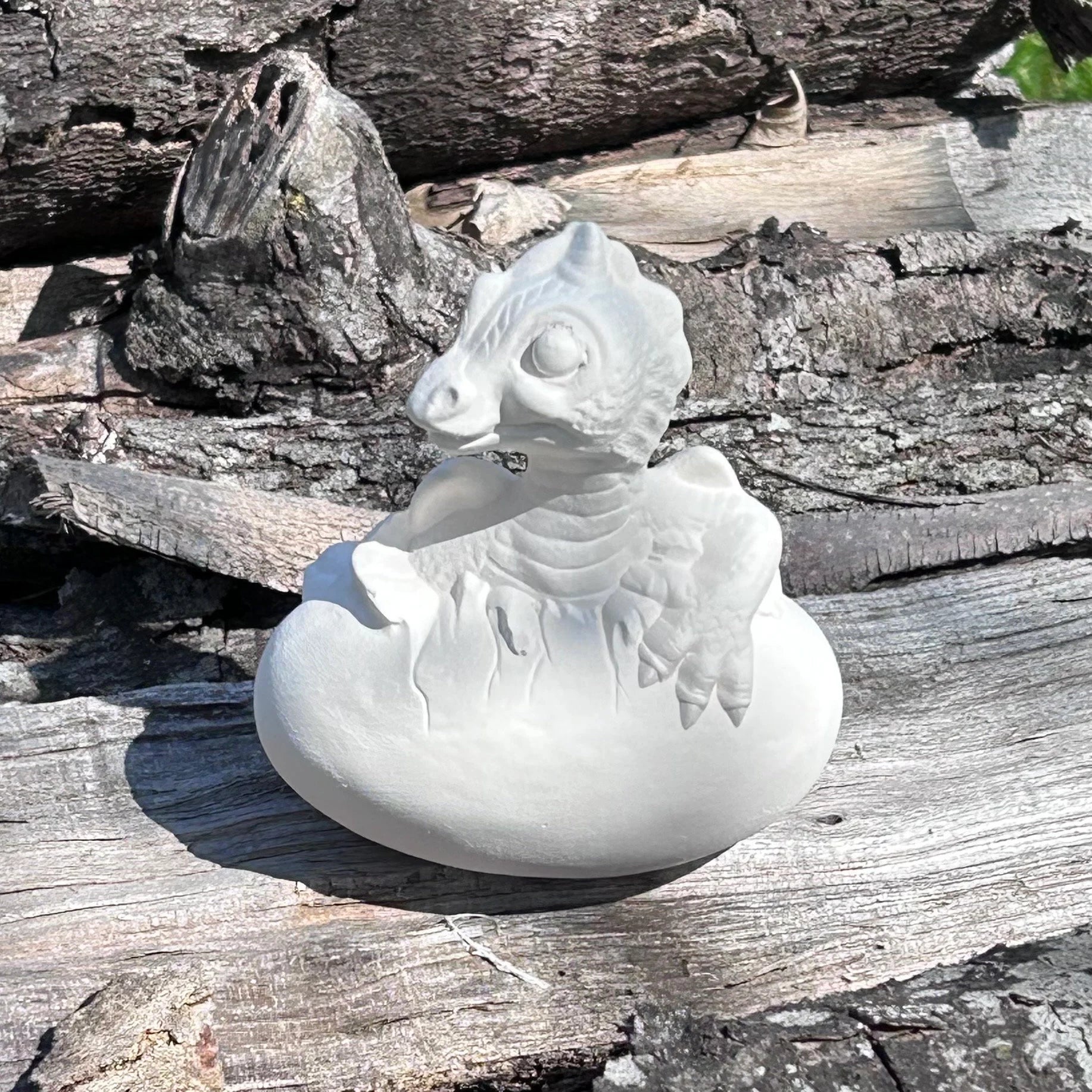 handmade ready to paint baby dragon and egg figurine sitting on a split log in the sunshine.