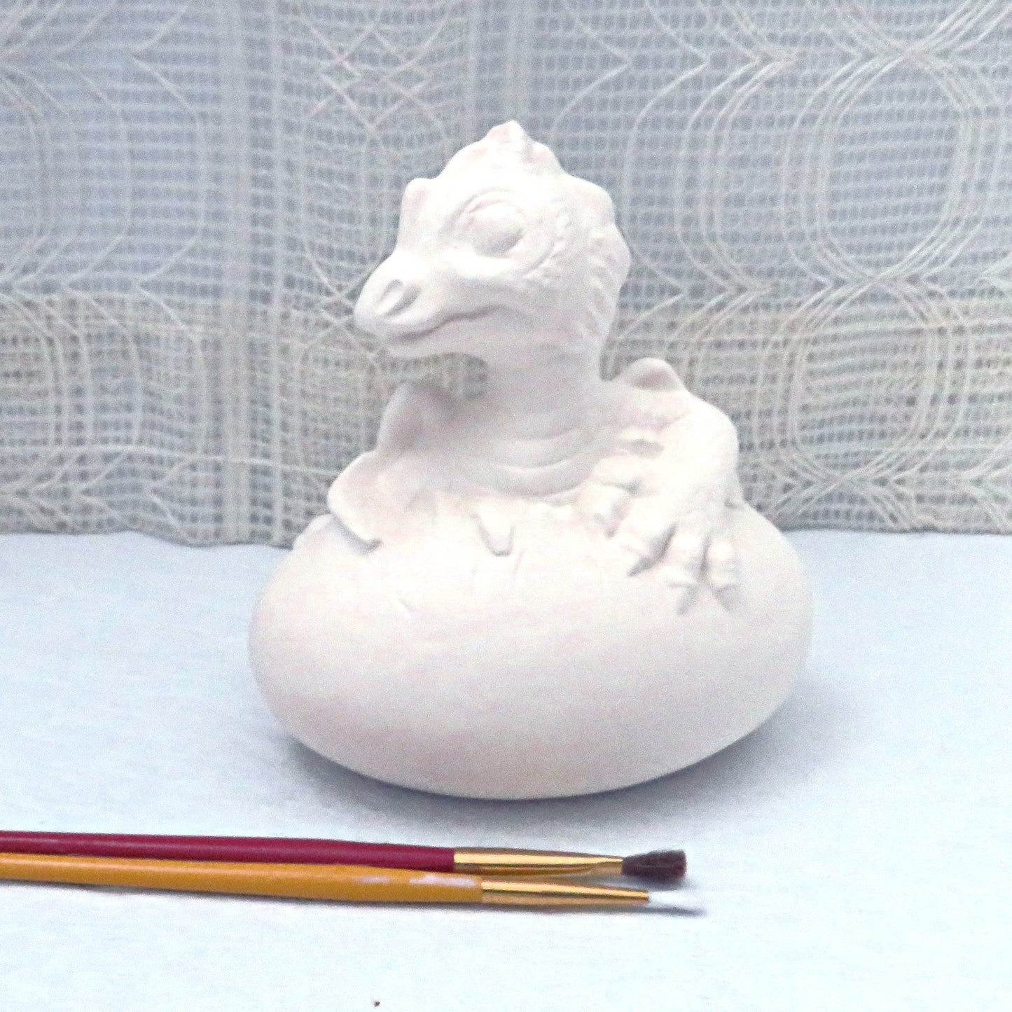 Ready to paint ceramic dragon figurine on a pale blue table with a lacy curtain behind it.  There are 2 paint brushes sitting in front of this dragon statue.
