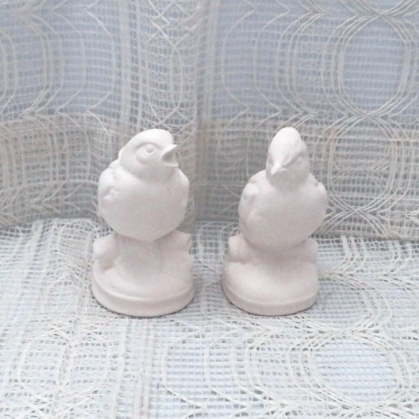 2 paintable ceramic bird figurines sitting on a lacy ecru table cloth with a lacy curtain behind them.