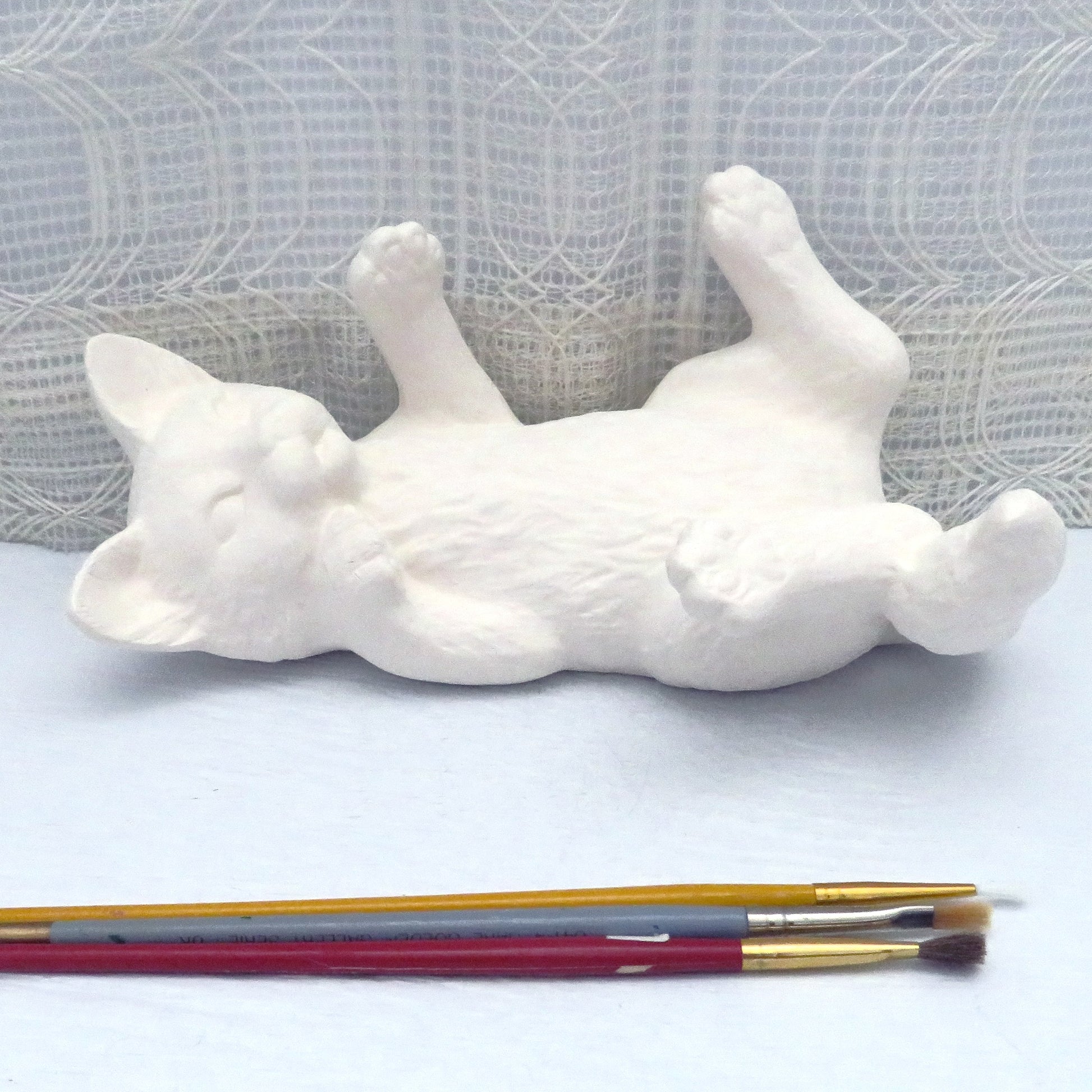 Ready to paint ceramic cat figurine positioned on his back with its legs in the air on a pale blue surface.  Facing camera.  Several paint brushes lying next to it.