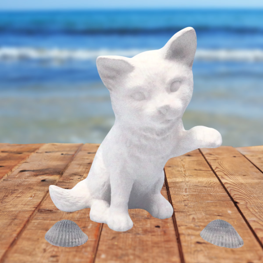 handmade unpainted ceramic cat figurine sitting with left paw up sitting on a table near the beach