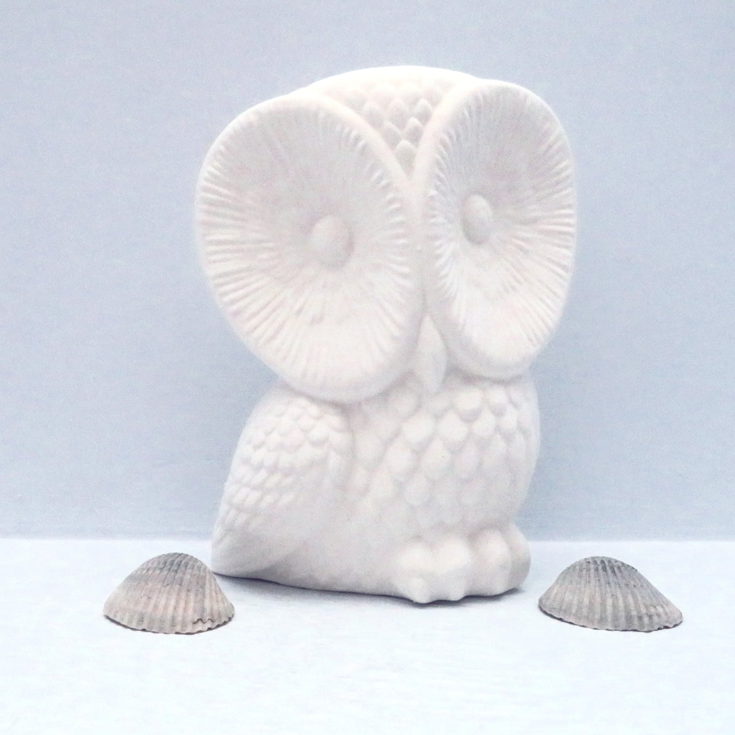 unpainted ceramic owl figurine on pale blue shelf with small sea shells next to it.