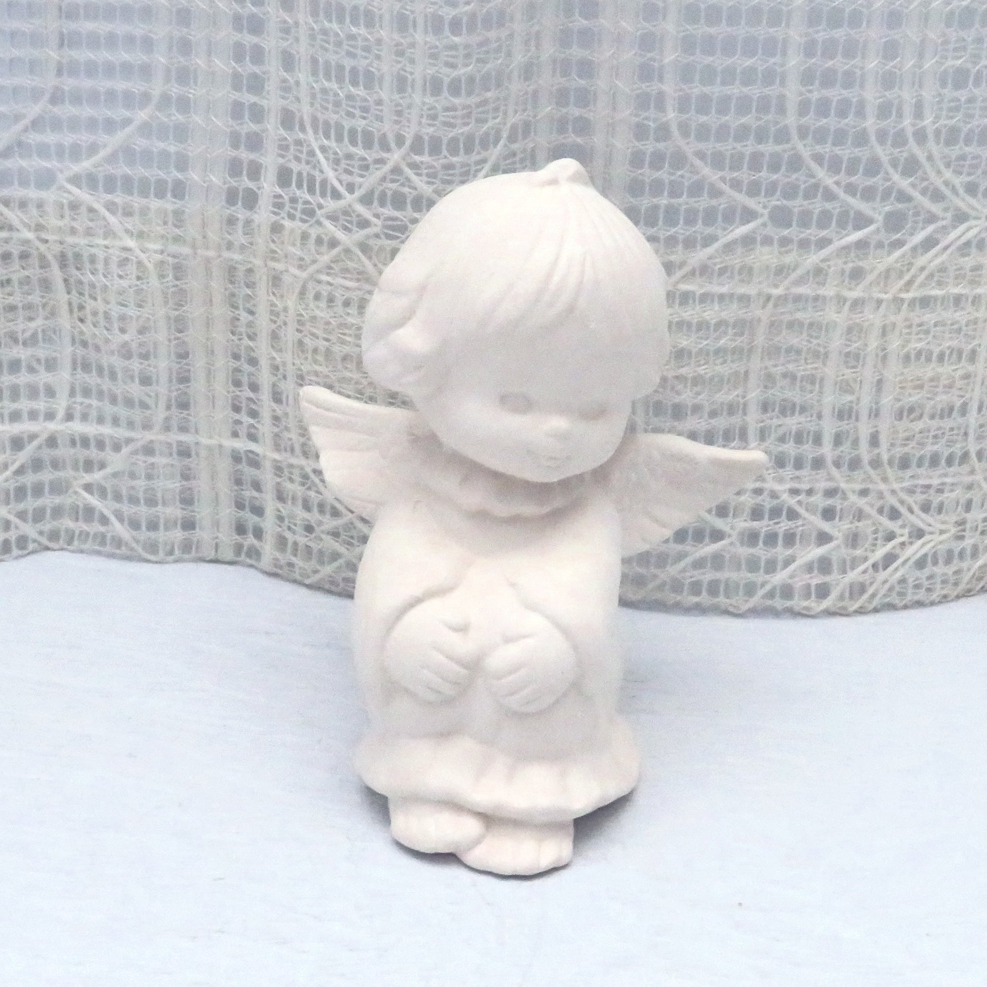 Handmade ready to paint ceramic angel sitting on pale blue table with ecru lacy curtain behind it.  Her wings are outstretched.  She is sitting with her knees up and her hands on her knees.