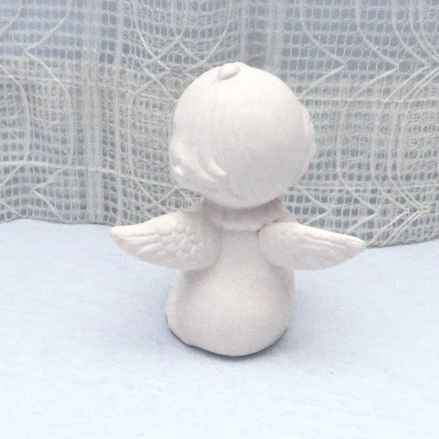 Rear view of unpainted ceramic angel showing wings and back of  head on a blue table.