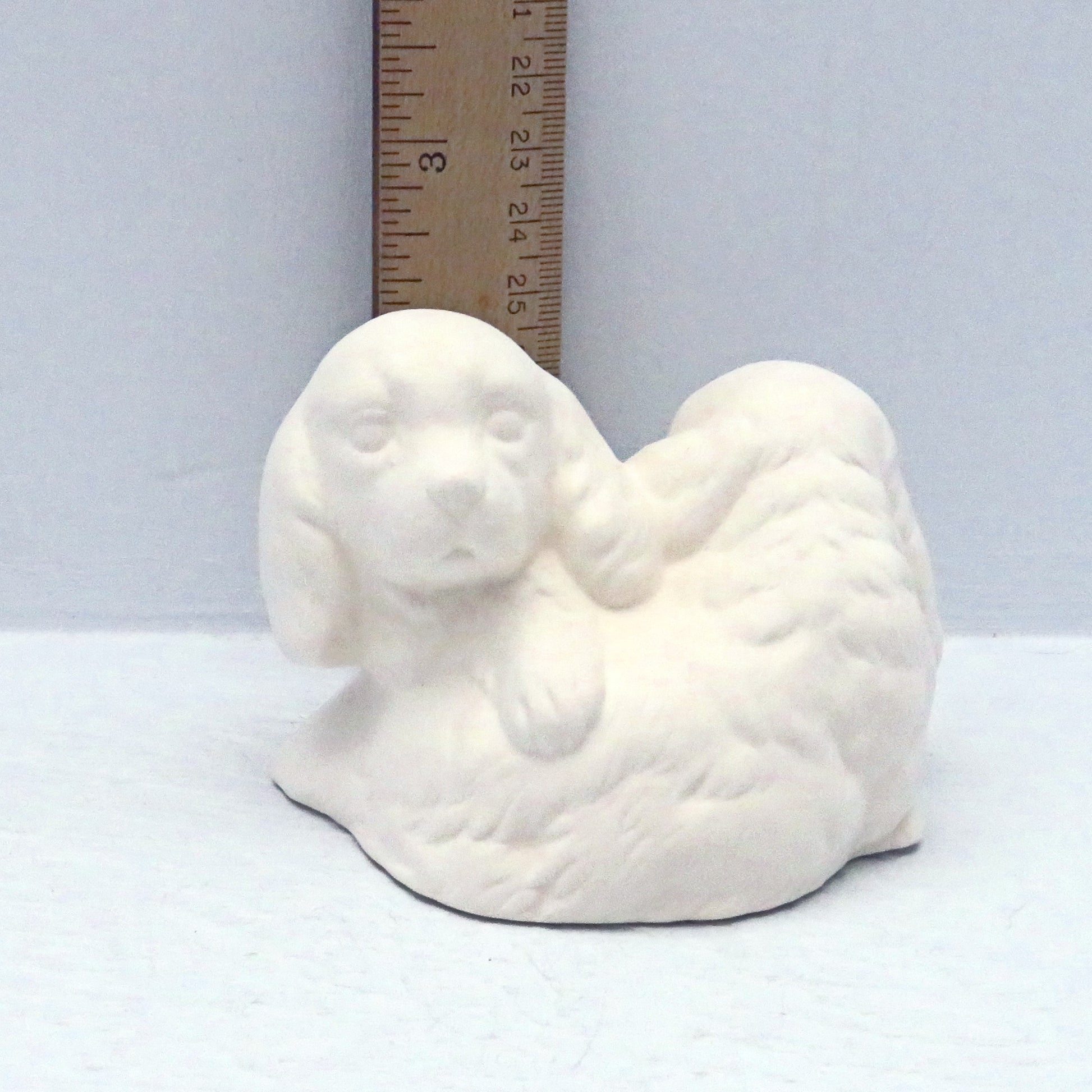 handmade ready to paint ceramic puppies statue near a ruler showing them to be approximately 2 1/4 inches tall against a pale blue background