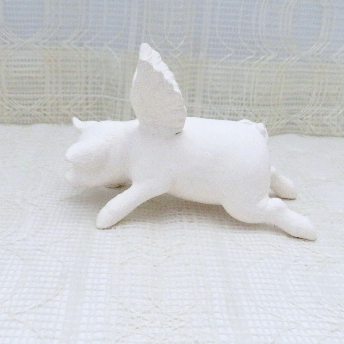 Handmade paintable ceramic flying pig angel on a lacy surface facing left.