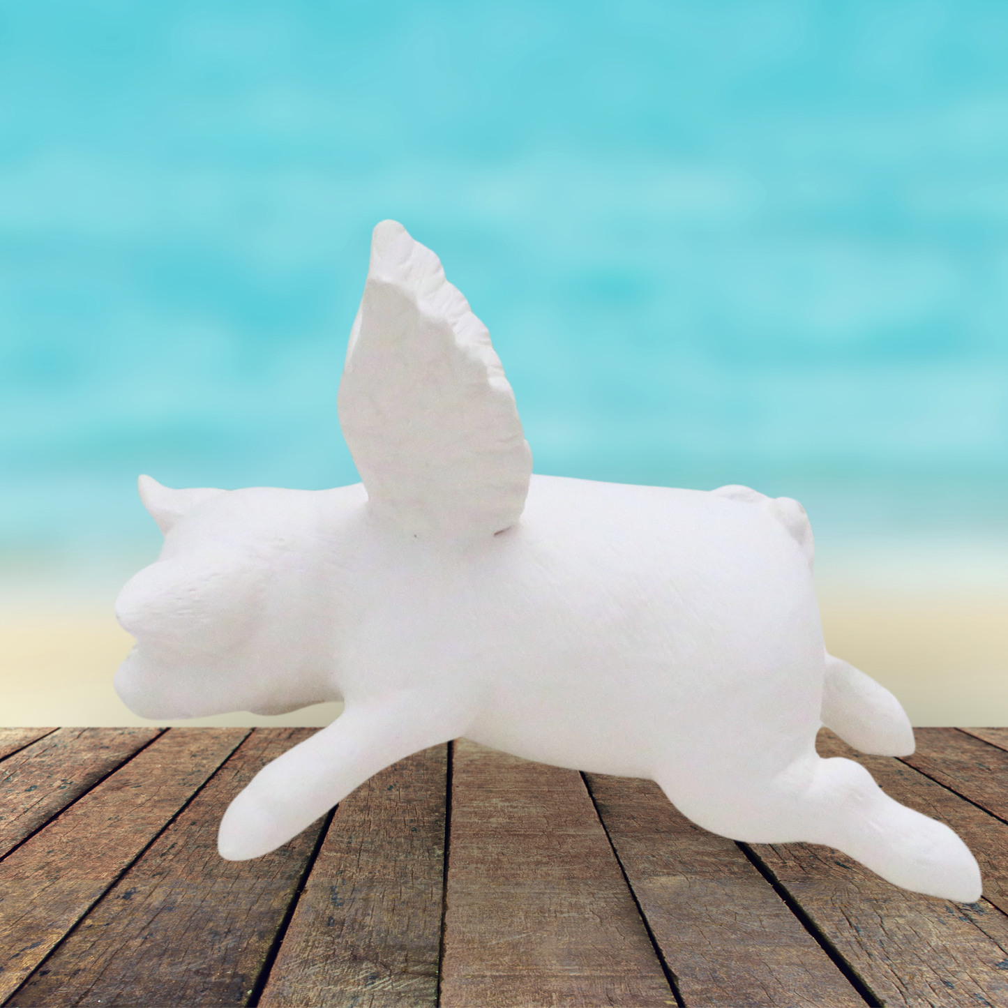 Handmade ready to paint ceramic flying pig angel on a rustic wood table near the ocean.