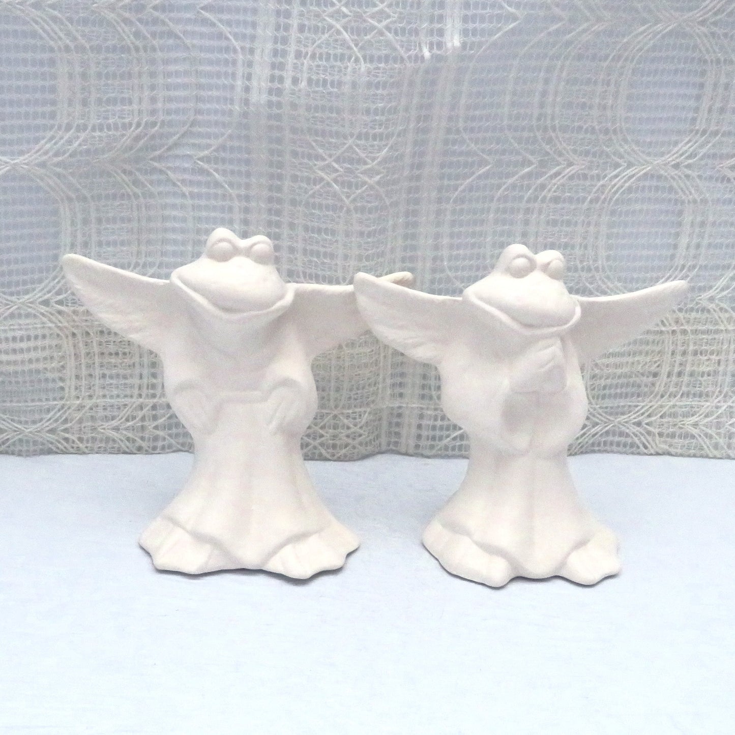 2 ready to paint ceramic frog angels standing on a pale blue table with an ecru lacy curtain behind them.  One has its hands at his sides.  The other has its hands in the prayer position.  The wings are outstretched.