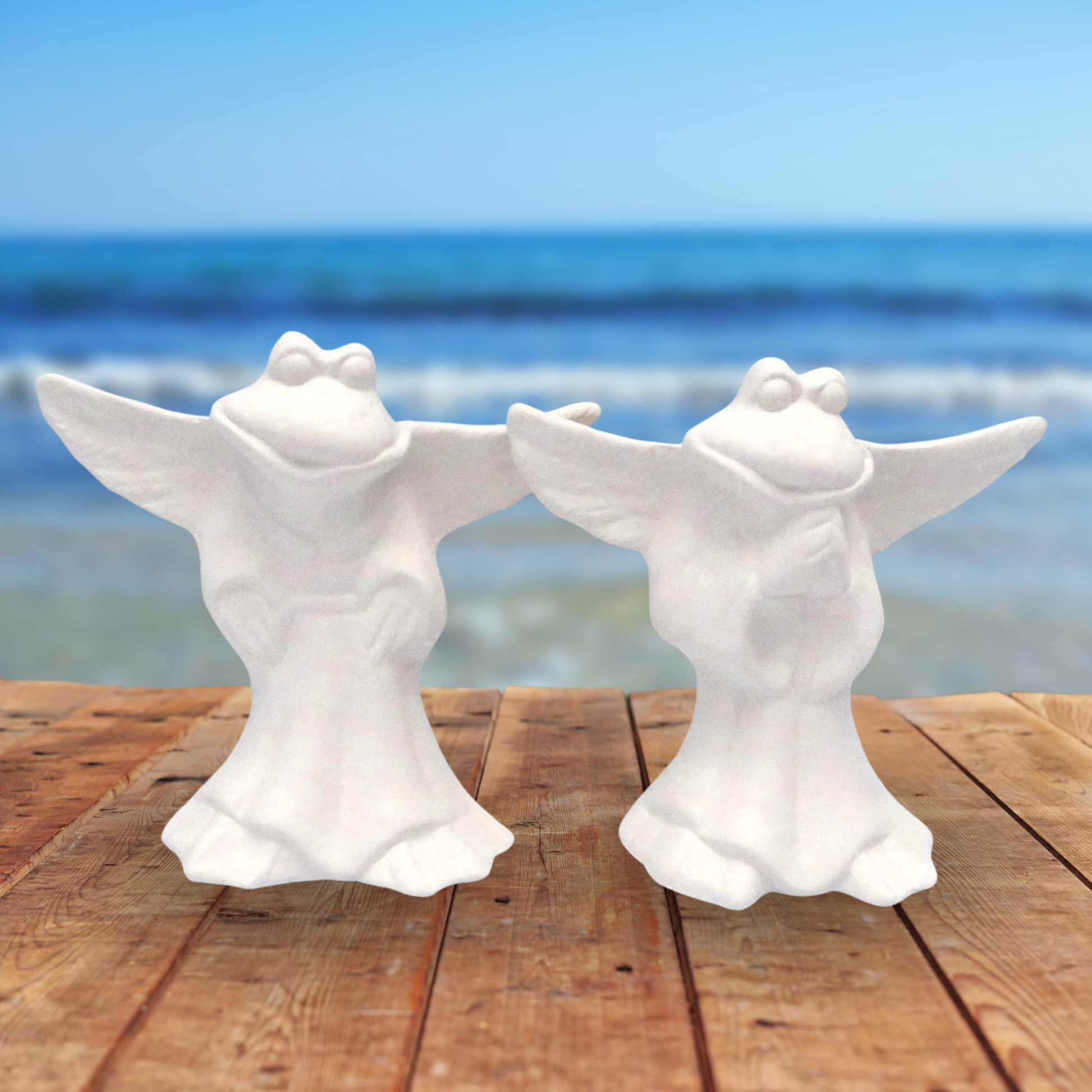 2 unpainted ceramic frog angels standing on a wood table with the ocean in the background.  One has its hands to the side.  The other has its hands in the prayer position.  The wings are outstretched.