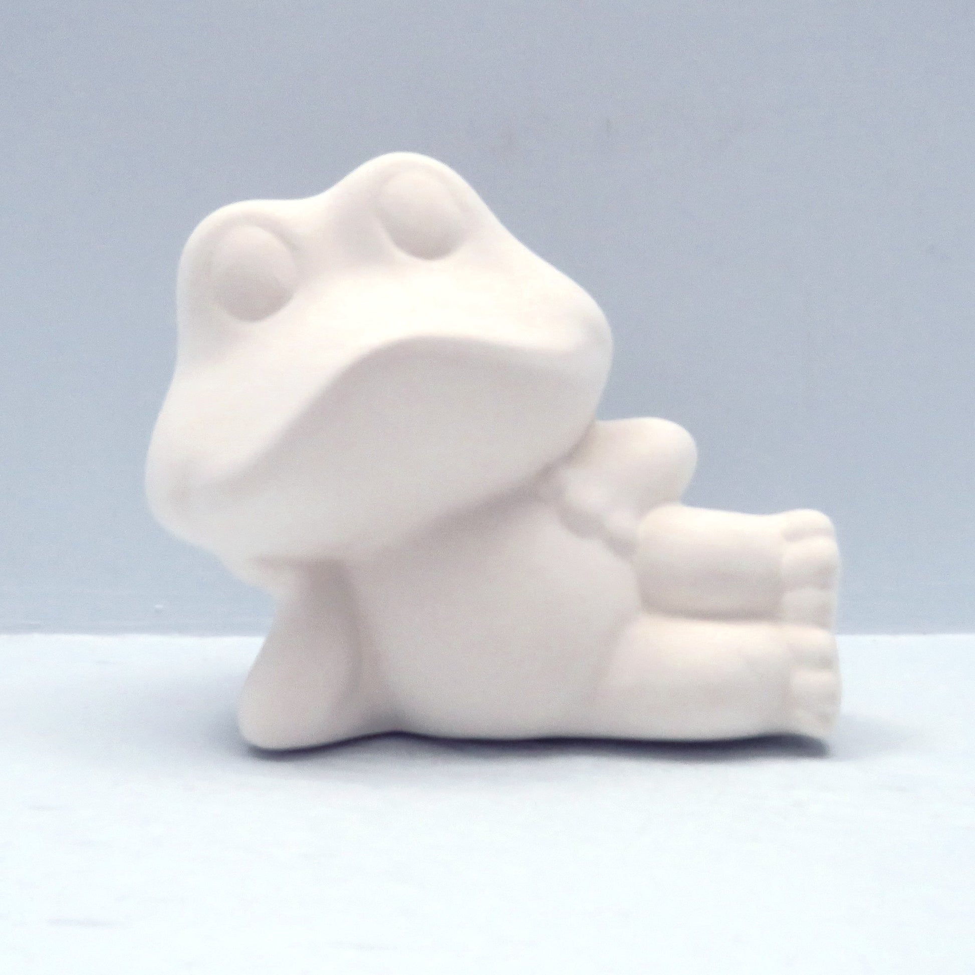 Paintable ceramic frog figurine lying on his right side with his head resting on his right hand on a blue background
