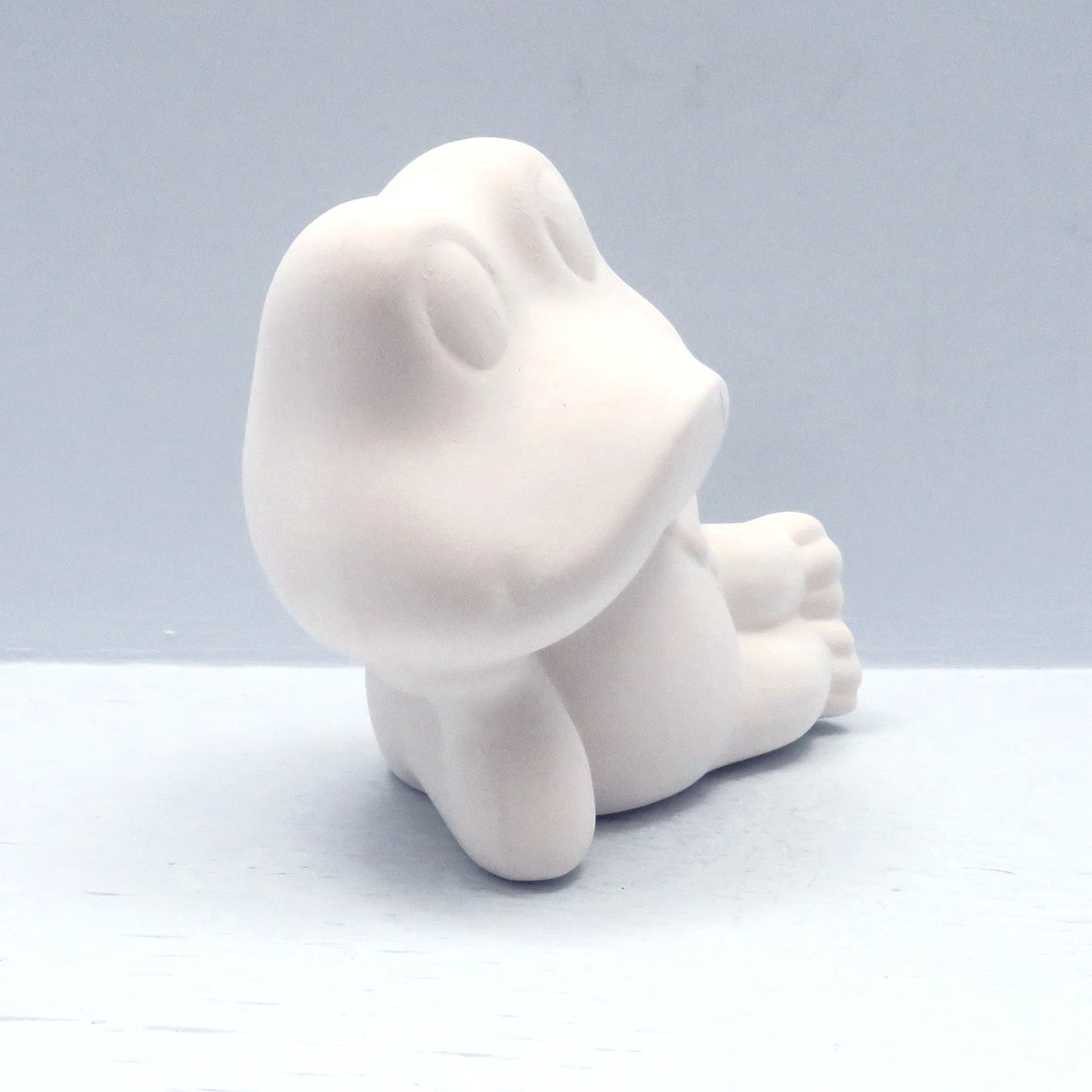 Ceramic frog figurine to paint lying on his side at an angle, so that his head is closest to the camera.