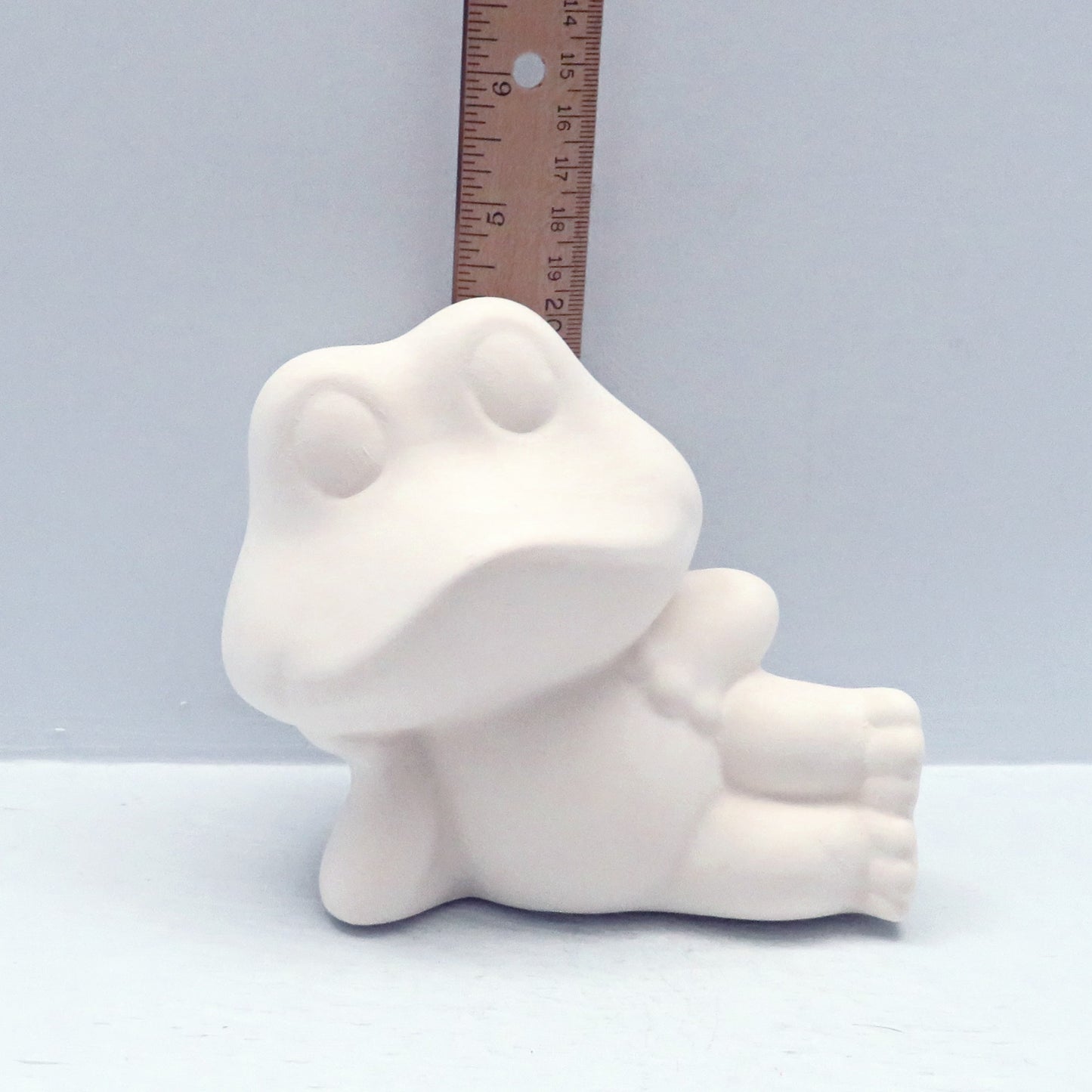 handmade ready to paint ceramic frog lying on his side near a ruler showing it to be just over 4 inches high