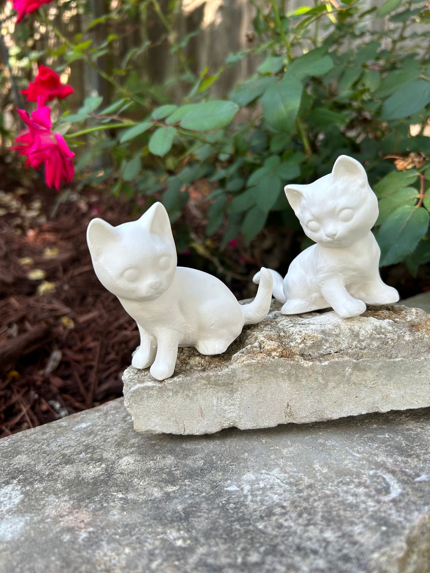 2 handmade sitting ready to paint cats sitting on white stones in front of a rose bush.  the cats are sitting wth their tails near each other and their faces forward