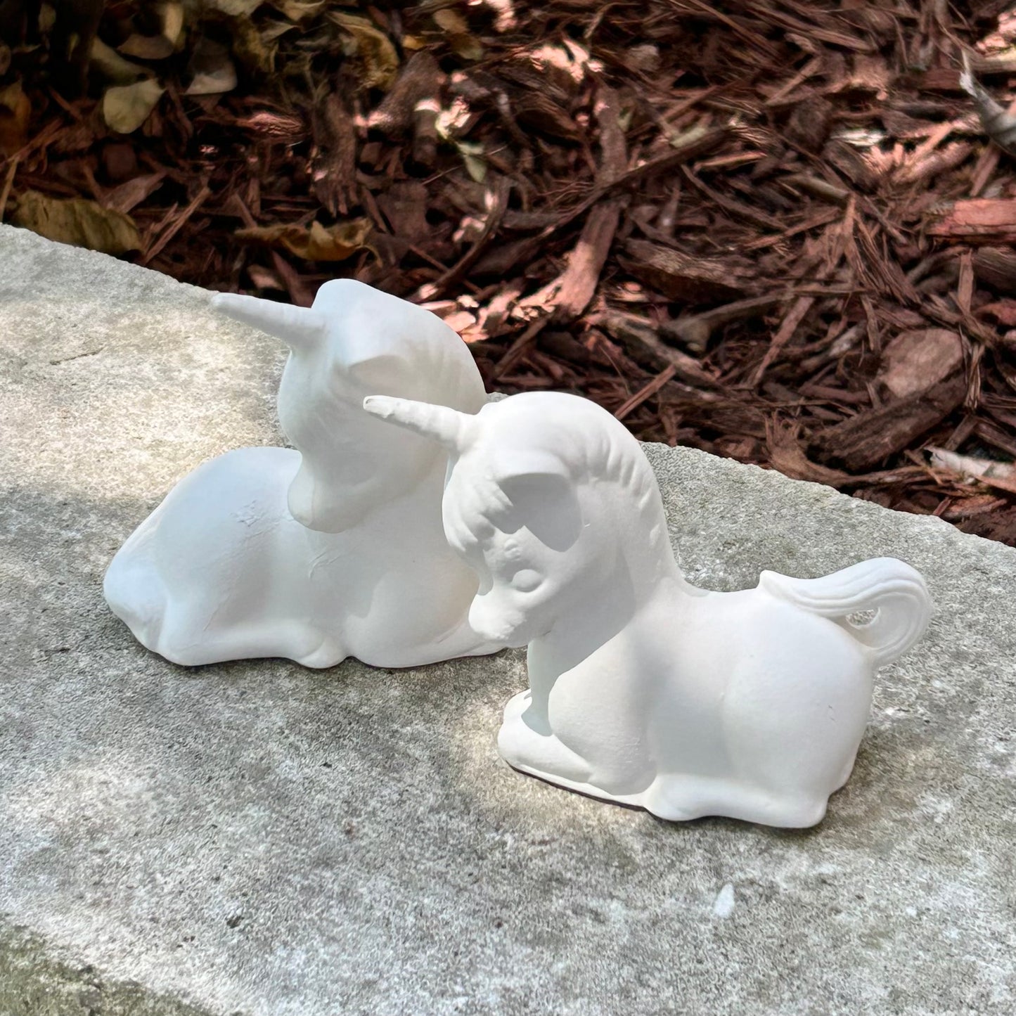 2 Ready to Paint Unicorn Figurines sitting on a white stone by a garden