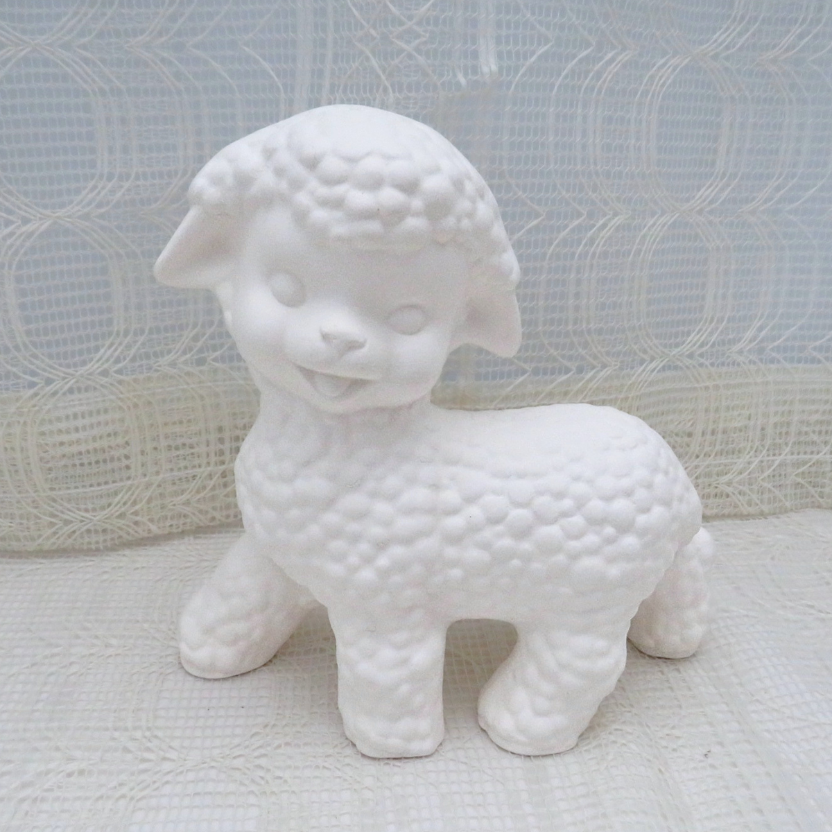 A white ceramic figurine of a cute lamb with textured wool, standing on a white patterned surface against a white lace curtain background.