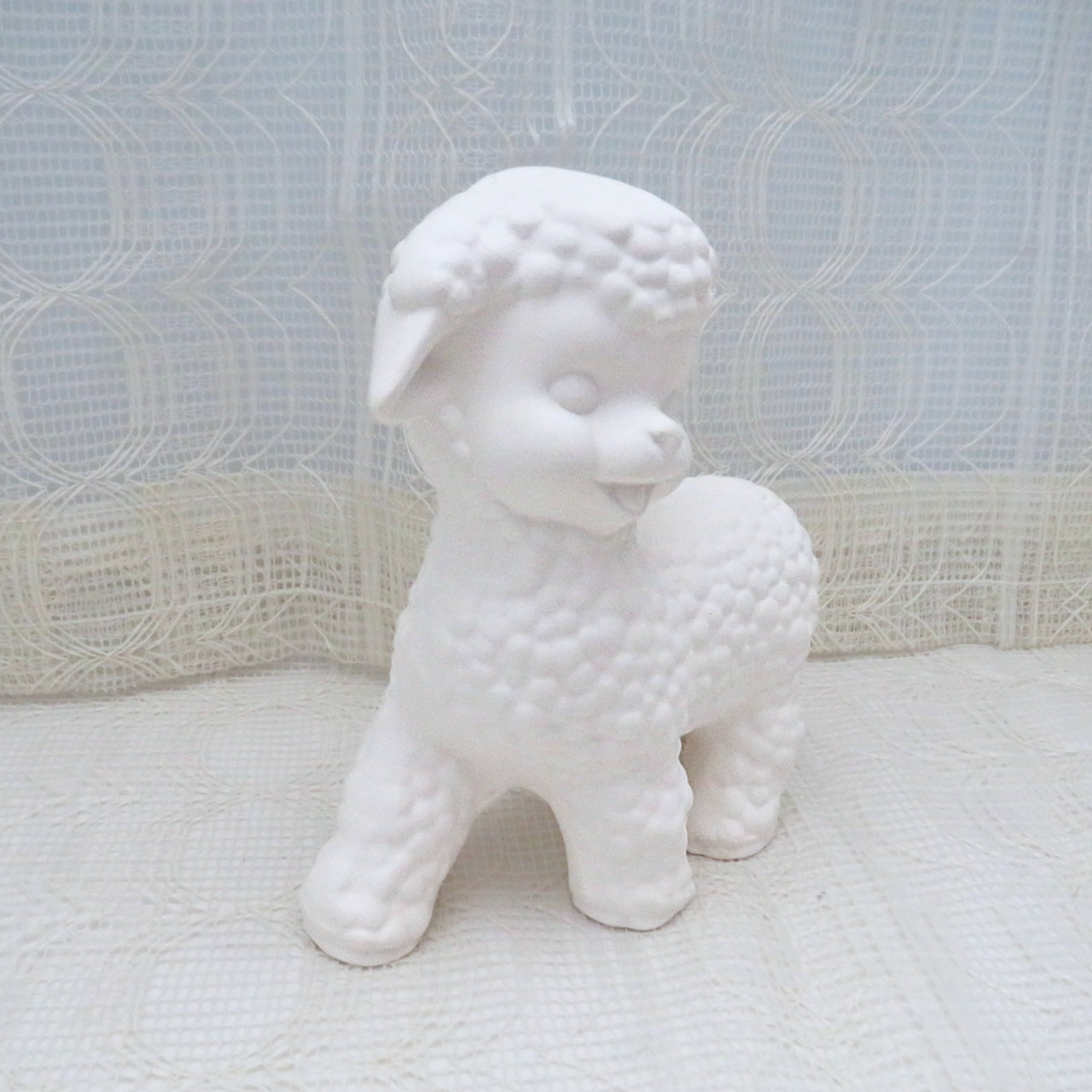 A white ceramic lamb figurine standing on a textured white surface with a white lace curtain backdrop.
