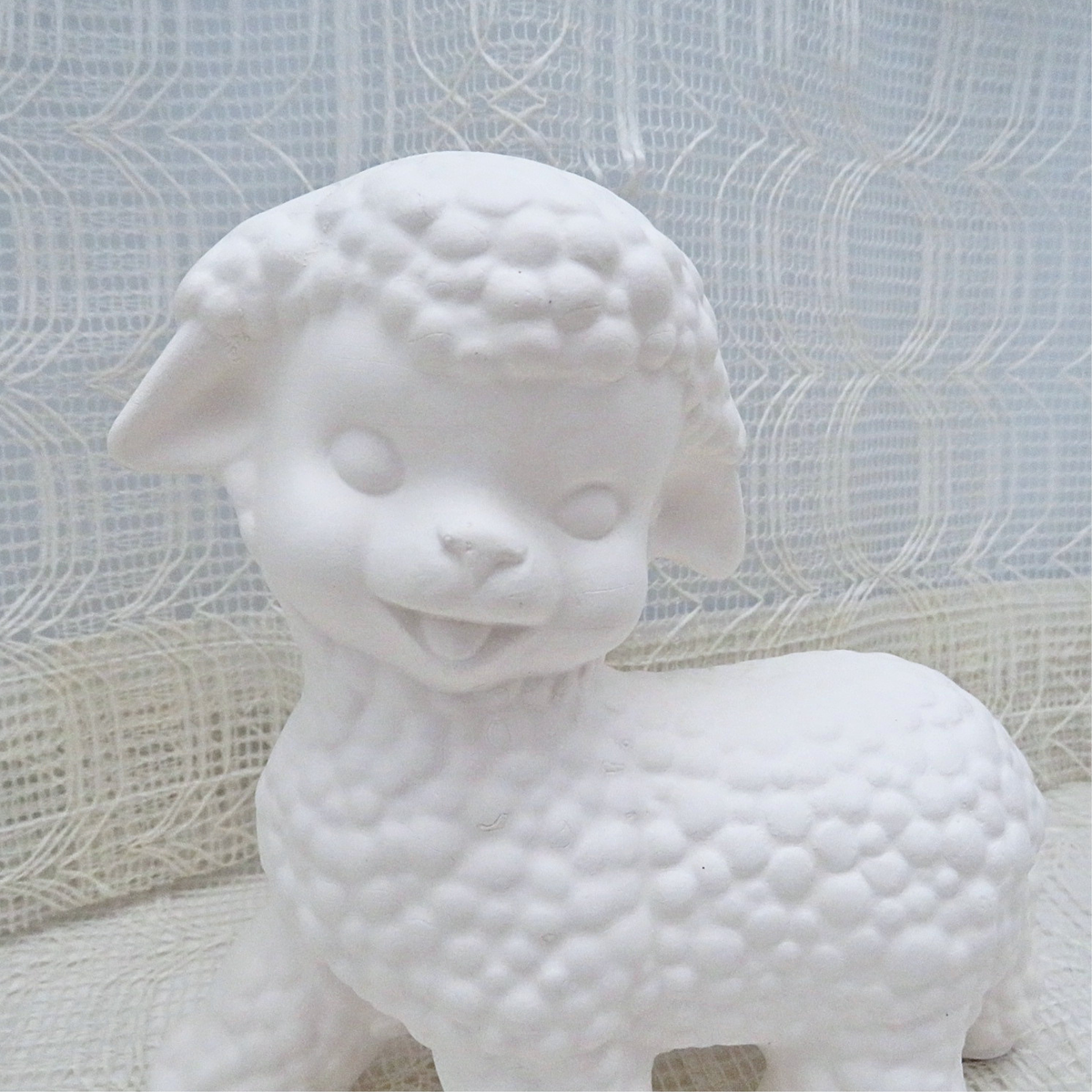 A unpainted ceramic lamb figurine with a textured, bubbly wool pattern, standing against a white lace curtain background.