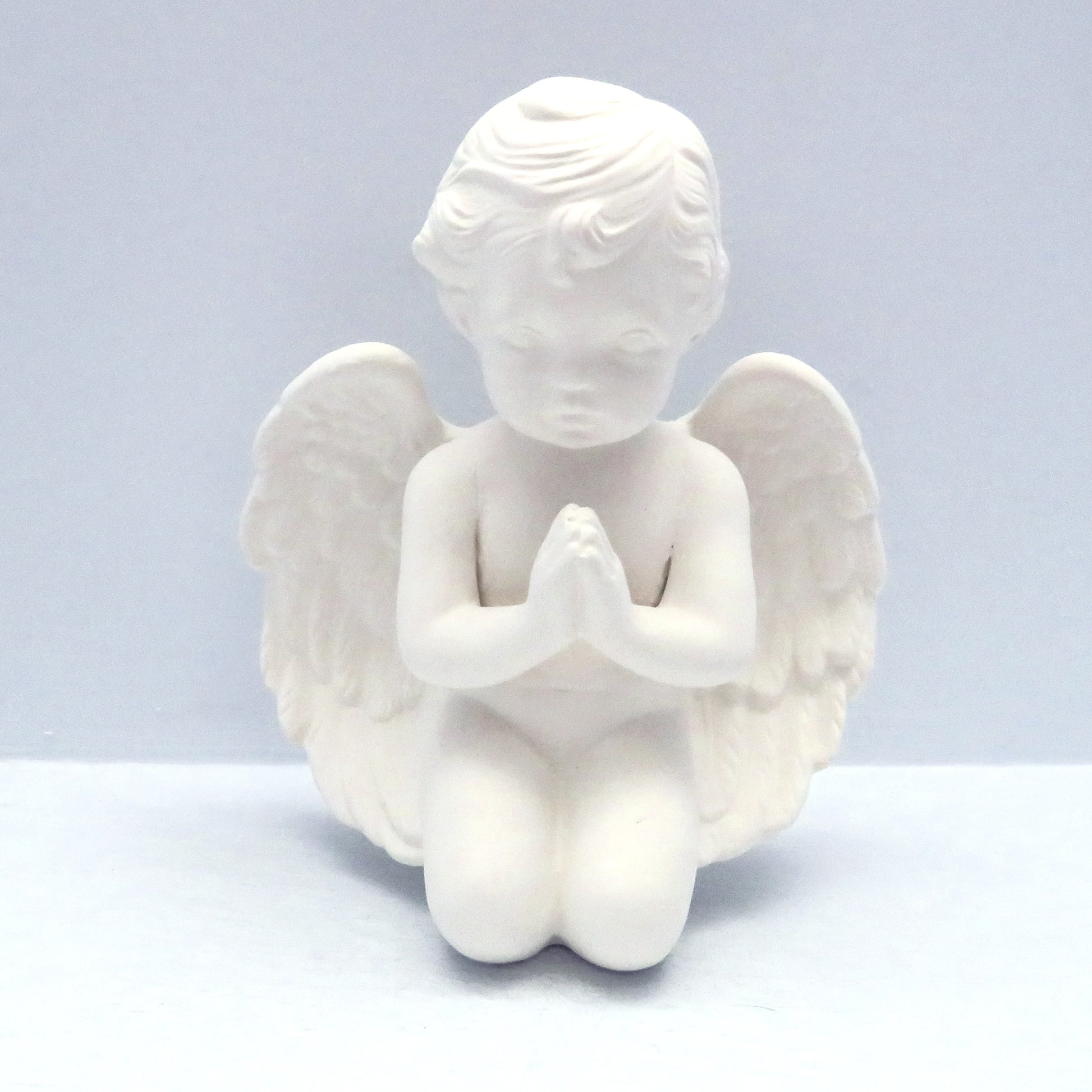 6 inch handmade ready to paint ceramic cherub angel figurine kneeling with his hands in the prayer position and its head bowed.  It is on a pale blue background