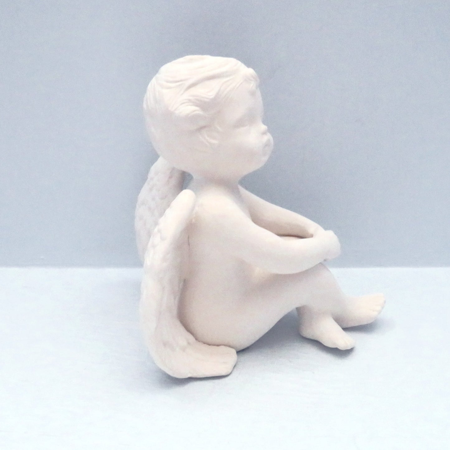 Side view of paintable ceramic cherub facing right.  His knees are bent and his legs are crossed at the ankles.  His elbows are bent, hands clasped and resting on his knees.  This angel statue is sitting on a pale blue table with pale blue background.