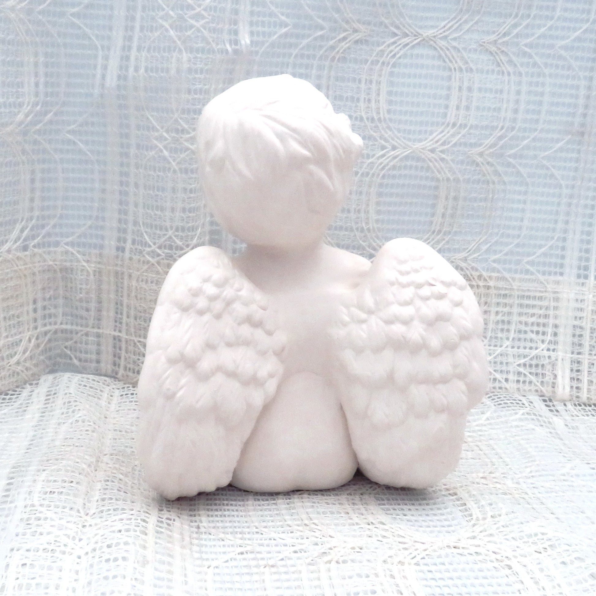 rear view focusing on wings of unpainted cherub figurine sitting on lacy ecru table cloth