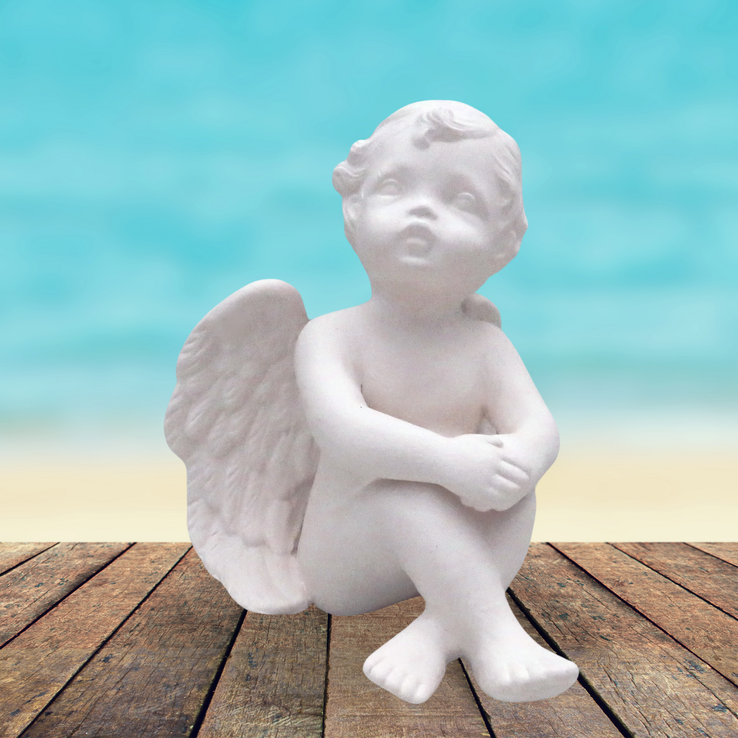 Large handmade ready to paint ceramic cherub with knees up sitting on a rustic wooden table near the ocean.