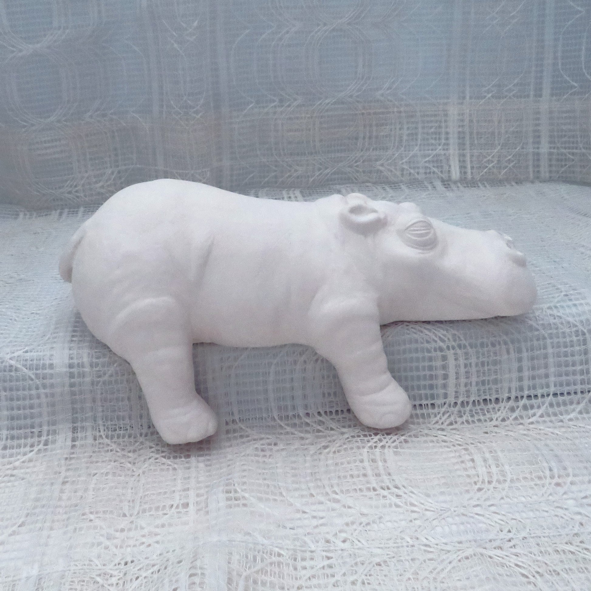 Large Ready to paint ceramic hippo figurine sitting on a shelf with its right legs hanging off.  There is an ecru lacy curtain behind and under it.